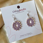 A pair of crocheted lavender, small, oval shaped beaded dangle earrings by Chirpyknots, displayed on a white brand card.