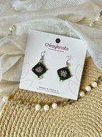 A pair of micro-crocheted granny square dangle earrings by Chirpyknots, featuring green outer and lavender center diamond-shaped crochet with silver hooks, displayed on a white brand card over a woven mat and white fabric and pearl beads.