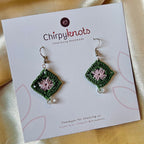 A pair of micro-crocheted granny square dangle earrings by Chirpyknots, featuring green outer and lavender center diamond-shaped crochet with silver hooks, displayed on a white brand card.