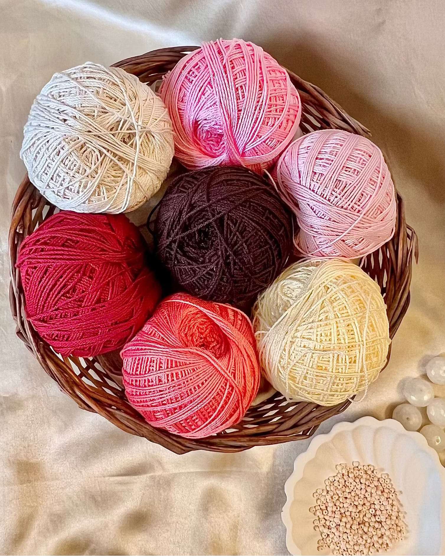 Six balls of variegated yarn in assorted colors arranged in a brown wicker basket on a beige satin fabric background, with a small white dish of seed beads nearby.